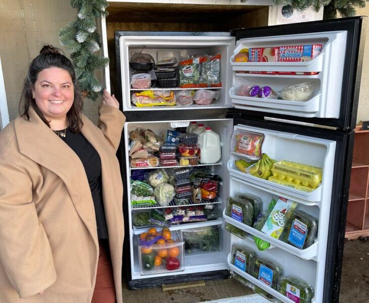 Picture of Executive Director, Shannon Cameron standing smiling at the camera in a brown long coat next to the Aurora Community Fridge with the fridge and freezer doors open displaying an abudance of food.